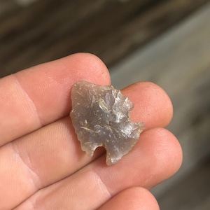 May include: A small, gray arrowhead with a chipped, irregular shape. The arrowhead is held in a hand, showcasing its size and texture. The stone appears translucent, with a slightly rough surface.
