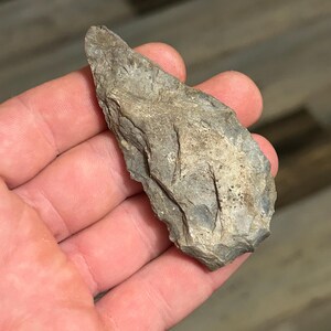 May include: A gray, chipped stone tool with a pointed end, held in a hand. The stone has a rough texture and appears to be an ancient artifact. The tool's shape suggests it may have been used for cutting or scraping.