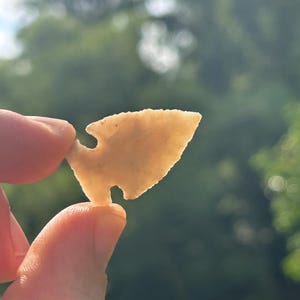 May include: A translucent, light beige stone arrowhead with a triangular blade, serrated edges, and a notched base. The object is held up to the light, revealing its smooth, chipped texture and distinct pointed tip and flared base shape.