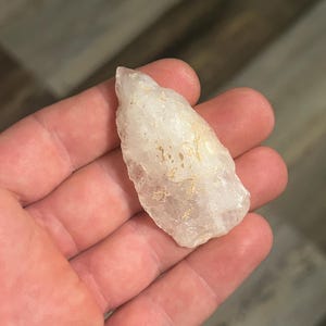 May include: A clear quartz crystal with a rough, natural shape. The crystal is held in a hand, showcasing its translucent quality and subtle variations in color. The crystal appears to be a raw, unpolished specimen.