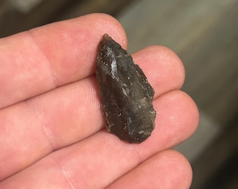 May include: A dark gray, chipped stone arrowhead with a sharp point. The arrowhead is held in a hand, showcasing its rough texture and shape. The stone appears to be a natural material, possibly flint or obsidian.