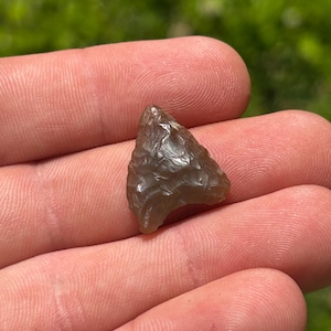 May include: A small, triangular arrowhead made of a translucent, gray-brown stone. The arrowhead has a chipped, textured surface and is held in a person's hand against a blurred green background.