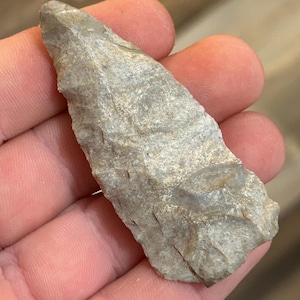 Puede incluir: Una punta de flecha de piedra gris con una punta puntiaguda y una base ligeramente curvada. La punta de flecha se sostiene en una mano.