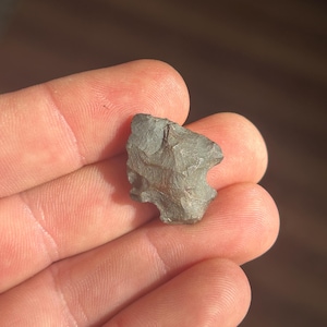 May include: A small, gray stone arrowhead with a rough, textured surface. The arrowhead is held in an open hand, showcasing its irregular shape and the natural variations in its color and texture. The arrowhead is a relic from a bygone era.