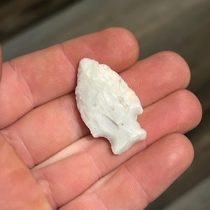 May include: A white arrowhead with a chipped, textured surface. The arrowhead is held in an open hand, showcasing its shape and detail. The stone artifact has a triangular shape with a pointed tip.
