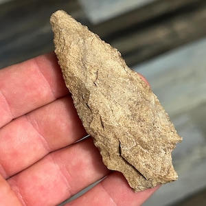 Puede incluir: Un artefacto de piedra de color beige claro, astillado, con una superficie rugosa y texturizada. La piedra tiene un extremo puntiagudo y parece ser un fragmento de un objeto más grande. El artículo se sostiene en una mano, mostrando su tamaño y forma.