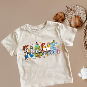 May include: Cream-colored t-shirt featuring the word "NAME" in colorful, cartoon-style letters. The letters are decorated with various characters and stars. A fun and playful design for kids.