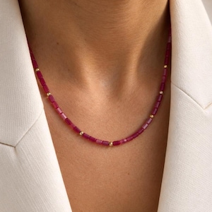 May include: A beaded necklace featuring small, rectangular, deep pink beads arranged in a continuous strand, interspersed with tiny, polished gold-toned spherical beads at regular intervals, resting against a white collared garment.