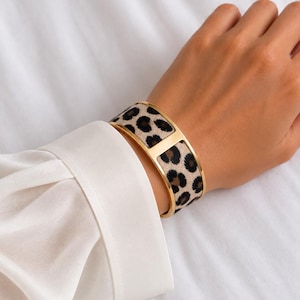 May include: A wide gold-tone metal cuff bracelet featuring a central horizontal bar and two curved panels inlaid with a tan and black leopard print pattern. The bracelet has a smooth, polished finish on the metal edges and a textured surface.