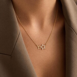 Op de afbeelding: Een goudkleurige ketting met twee stervormige hangers. De ene ster is een eenvoudige open gouden omtrek, terwijl de aangrenzende ster iets groter is en over het gehele oppervlak is ingelegd met kleine, heldere, fonkelende steentjes.