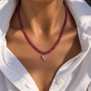 Pode incluir: Um colar com um fio de pequenas contas redondas em magenta profundo. Um único pendente facetado em forma de coração cor-de-rosa claro está suspenso no centro do fio de contas por um pequeno elo de metal dourado sobre uma camisa branca.