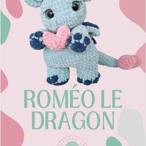May include: A crocheted blue dragon holding a pink heart. The dragon has a pink horn and blue feet. The text "ROMEO LE DRAGON" is written in green letters below the dragon. The text "PATRON CRÉÉ PAR @CAPTAINE CRO ! TOUS DROITS RÉSERVÉS" is written in black letters below the text.