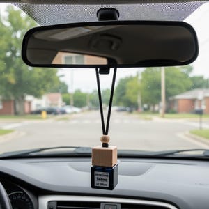 Könnte beinhalten: Ein Auto-Lufterfrischer, der am Rückspiegel hängt. Der Lufterfrischer hat einen Holzquader auf einer dunkelblauen quadratischen Flasche. Die Flasche hat ein weißes Etikett mit dem Text "Balance Car Diffuser". Der Hintergrund zeigt eine Wohnstraße.