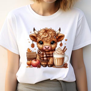 May include: White t-shirt featuring a cartoon Highland cow with a brown and white plaid scarf. The cow is next to a mug of coffee with whipped cream, an apple, and a small wooden sign. Autumn-themed design.