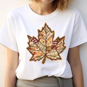 May include: White t-shirt featuring a large, detailed maple leaf design. The leaf is composed of various plaid and floral patterns in autumn colors like orange, red, and green, with a brown outline. The shirt is a casual, everyday wear item.