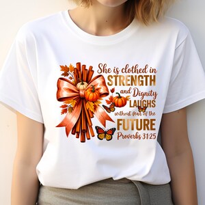 May include: White t-shirt with a fall-themed design. The graphic includes an orange bow, pumpkins, autumn leaves, and butterflies. Text reads "She is clothed in STRENGTH and Dignity LAUGHS without fear of the FUTURE Proverbs 31:25."