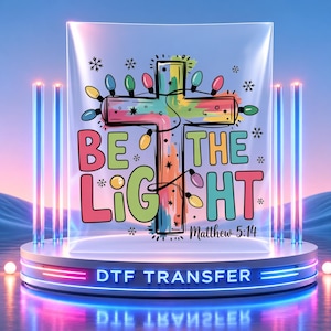 Peut inclure: Une feuille de transfert DTF présentant une croix colorée décorée de guirlandes lumineuses multicolores et de petits flocons de neige noirs. Le texte indique BE THE LIGHT et Matthew 5:14 dans une police audacieuse. Couleurs rose, bleu et vert.