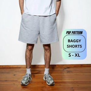 May include: Light grey baggy shorts paired with a plain white t-shirt, white socks, and grey athletic trainers. A digital graphic overlay on the right side displays the text: PDF PATTERN BAGGY SHORTS S - XL within a rounded rectangular border.