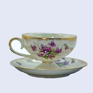 May include: A white teacup and saucer set with a gold rim and a floral design featuring purple flowers. The flowers are painted in a realistic style and have green leaves.