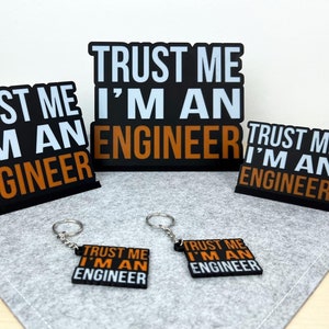 Op de afbeelding: Vier zwarte en oranje borden met de tekst "Trust me I'm an engineer" op een grijs oppervlak. Twee van de borden zijn groter en twee zijn kleiner. De kleinere borden zijn bevestigd aan sleutelhangers.