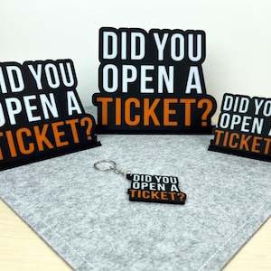 Könnte beinhalten: Vier schwarze und orangefarbene Schilder mit dem Text "Haben Sie ein Ticket geöffnet?" darauf. Die Schilder sind in verschiedenen Größen und werden auf einer grauen Oberfläche angezeigt. Eines der Schilder ist an einem silbernen Schlüsselanhänger befestigt.