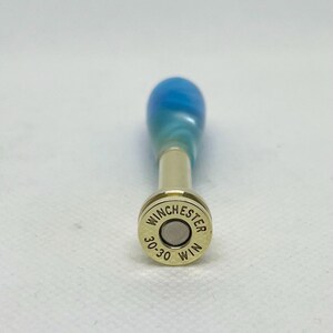 May include: A bottle stopper with a blue and teal marbled handle and a gold-colored metal base. The base is a Winchester 30-30 WIN bullet casing.