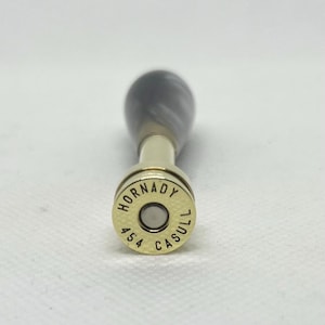May include: A bottle stopper with a gold-colored base and a dark gray and white marbled handle. The base is engraved with "HORNADY" and "454 CASULL".