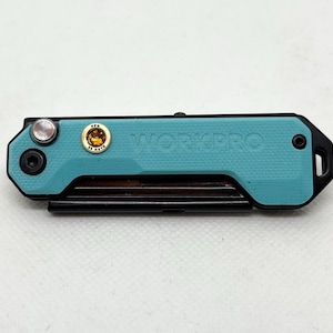 May include: A teal multi-tool with the word "WORKPRO" printed on the side. The tool has a black frame and a small gold emblem. It features a silver button and a black loop on one end. The tool is designed for various tasks.