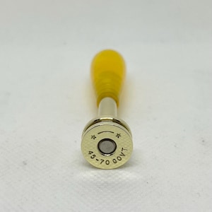May include: A bottle stopper with a yellow handle and a silver metal base. The base is engraved with the text "45-70 GOVT" and star symbols. The stopper is designed to fit into a bottle.