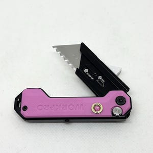 May include: A pink WORKPRO utility knife with a black blade holder. The blade is partially extended, revealing a serrated edge. The knife has a small, decorative jewel and a silver button. The text "PUSH UP" and "PULL" are visible.