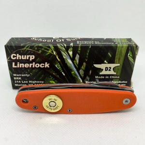 May include: An orange Chirp Linerlock knife with a D2 blade, featuring a brass accent with "W-W 45-70 GOVT" text. The knife is set in front of its box, which has the text "School of Sun" and "Designed by Randall's Adventure & Training".