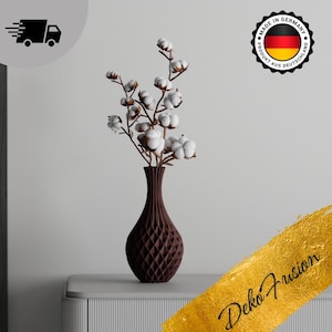 Könnte beinhalten: Eine braune, strukturierte Keramikvase mit geometrischem Muster hält einen Strauß weißer Baumwollblumen. Die Vase steht auf einer grauen Oberfläche mit goldenem Hintergrund.
