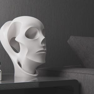 Könnte beinhalten: Eine weiße, abstrakte Skulptur eines menschlichen Kopfes mit großen Augenhöhlen und einem stilisierten Ohr. Die Skulptur steht auf einer dunklen Oberfläche, mit einer grauen Wand und einem grauen Sofa im Hintergrund. Modernes, minimalistisches Design.