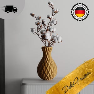 Könnte beinhalten: Eine goldene, geometrische Vase mit strukturierter Oberfläche hält einen Strauß weißer Baumwollstängel. Die Vase steht auf einer grauen Oberfläche mit goldenem Hintergrund.