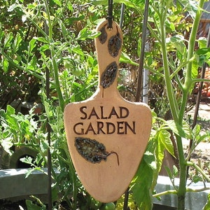 May include: A brown ceramic garden marker with the words "Salad Garden" written on it. The marker is hanging from a metal stake in a garden.