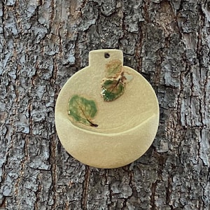 May include: A circular, beige ceramic wall planter with a flat top featuring a small hole for hanging. The front surface is decorated with two small, green, textured leaf-shaped accents. The planter has a curved, open pocket design at the bottom.
