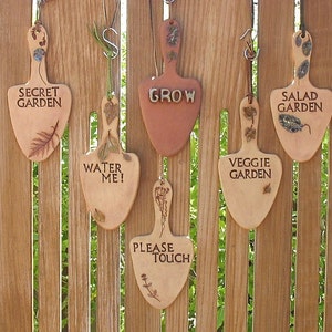 May include: Six wooden garden signs shaped like trowels, hanging on a wooden fence. The signs are decorated with dried flowers and leaves. The signs read: "Secret Garden", "Water Me!", "Grow", "Veggie Garden", "Salad Garden", and "Please Touch".