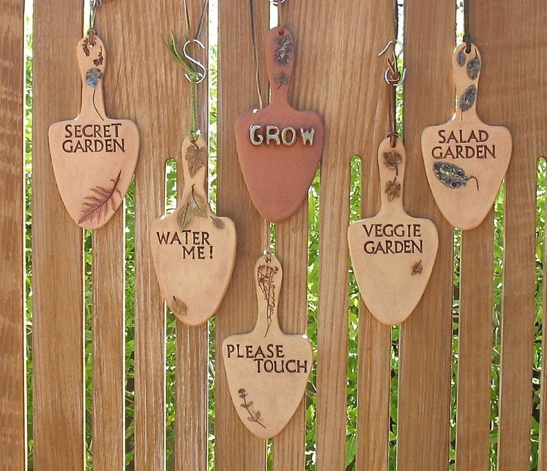May include: Six wooden garden signs shaped like trowels, hanging on a wooden fence. The signs are decorated with dried flowers and leaves. The signs read: "Secret Garden", "Water Me!", "Grow", "Veggie Garden", "Salad Garden", and "Please Touch".