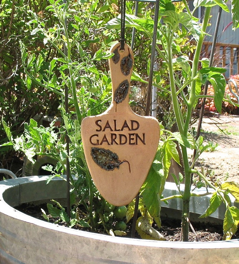 May include: A brown ceramic garden marker with the words "Salad Garden" written on it. The marker is hanging from a metal stake in a garden.