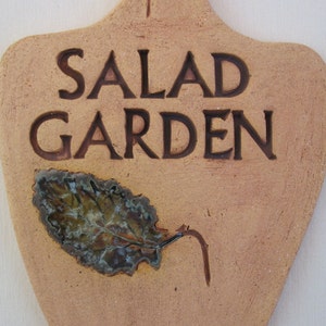 May include: A terracotta-colored ceramic sign with the words "SALAD GARDEN" in brown lettering. A stylized leaf with a green and brown glaze is attached to the sign.
