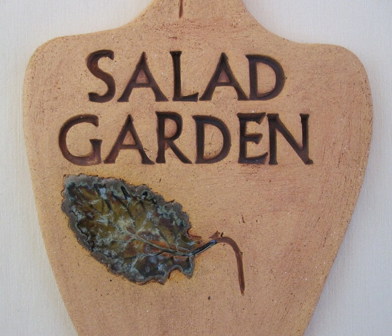 May include: A terracotta-colored ceramic sign with the words "SALAD GARDEN" in brown lettering. A stylized leaf with a green and brown glaze is attached to the sign.