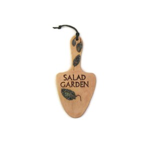 May include: A light brown wooden sign shaped like a spade with the text "SALAD GARDEN" in brown lettering. The sign has a black string attached to the top and is decorated with dried leaves.