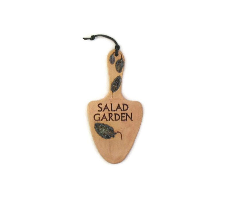 May include: A light brown wooden sign shaped like a spade with the text "SALAD GARDEN" in brown lettering. The sign has a black string attached to the top and is decorated with dried leaves.