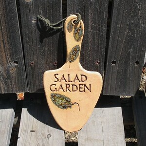 May include: A ceramic garden marker shaped like a spade with the words "SALAD GARDEN" in brown lettering. The marker is decorated with green and brown leaf designs.