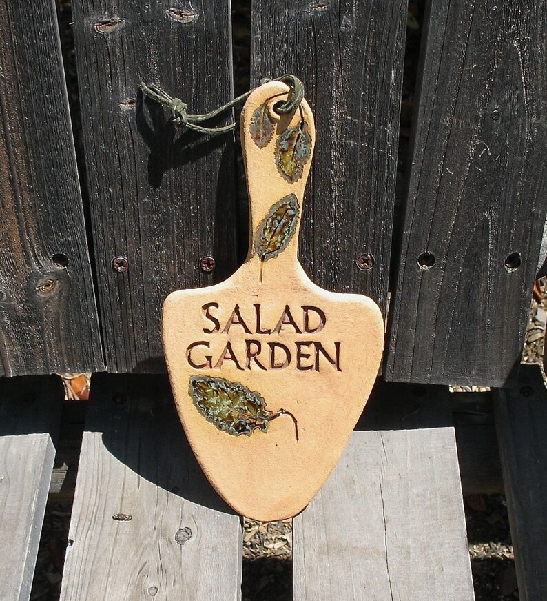 May include: A ceramic garden marker shaped like a spade with the words "SALAD GARDEN" in brown lettering. The marker is decorated with green and brown leaf designs.