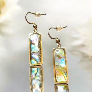 May include: A pair of gold-coloured rectangular drop earrings. Each earring has a shimmering, iridescent abalone shell inlay. The earrings have a simple hook design for pierced ears. The background is blurred, with white flowers.