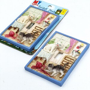 May include: Two decorative light switch plates with a cat theme. Each plate features a blue frame and a detailed scene with a white cat, a gray cat, a window, and a ball of yarn. The plates are new and still in their original packaging.