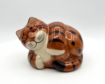 Peut inclure: Figurine en céramique représentant un chat endormi. Le chat est brun avec des rayures et des taches noires, et a un ventre et un visage blancs. Le chat est recroquevillé dans une pose détendue, les yeux fermés.