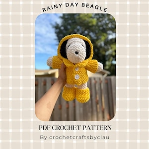 Op de afbeelding: Een gehaakte beagle knuffel die een felgele regenjas met capuchon draagt, voorzien van twee witte knopen en bijpassende gele regenlaarzen. De tekst RAINY DAY BEAGLE, PDF CROCHET PATTERN en By crochetcraftsbyclau staat op de afbeelding.