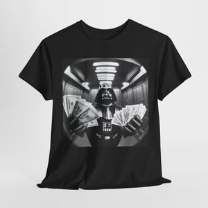 May include: A black and white photo of Darth Vader holding stacks of money in each hand. The photo is printed on a black t-shirt.
